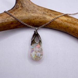 May include: A silver necklace with a teardrop-shaped pendant. The pendant features a silver cap and is filled with iridescent, white, and pastel-colored stones. The necklace is displayed on a light brown antler against a white background.
