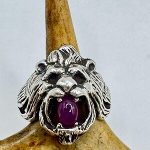 May include: A silver lion head ring with a purple gemstone set in the mouth.