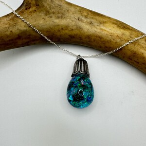 May include: A silver chain necklace with a pendant. The pendant is a teardrop shape with a silver cap and a blue and green speckled design. The necklace is displayed on a light background with a brown antler.