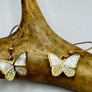 May include: Gold-tone butterfly earrings with white iridescent wings and a lower section encrusted with small, clear crystals. The earrings are displayed against a brown, textured background.