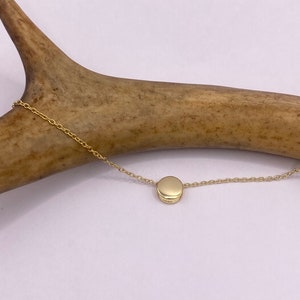 Puede incluir: Un collar de cadena de oro con un pequeño colgante redondo. El colgante es liso y tiene una forma ligeramente ovalada.