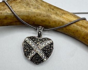 Puede incluir: Un collar de plata con un colgante en forma de corazón con un diseño en blanco y negro. El colgante está adornado con pequeñas piedras negras y brillantes transparentes.
