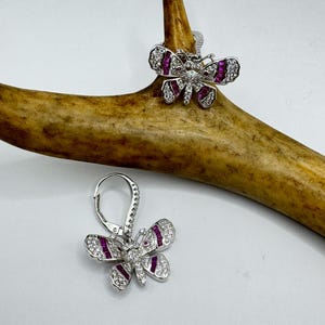 May include: A pair of silver butterfly earrings with clear and purple stones. The earrings are hanging from a brown antler.