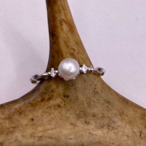 May include: A silver ring with a single white pearl and small clear stones set in the band.