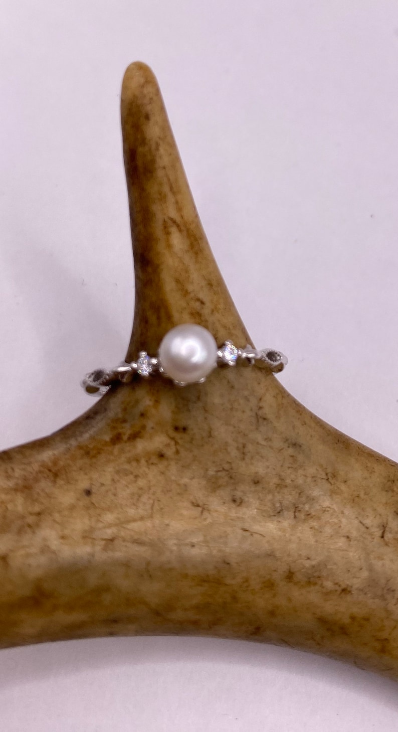 May include: A silver ring with a single white pearl and small clear stones set in the band.