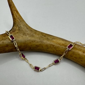 Op de afbeelding: Een gouden kettingarmband met vijf rechthoekige robijnrode stenen.