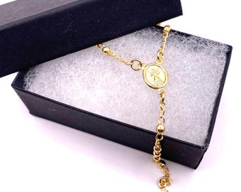 May include: A gold-coloured rosary with a small cross charm and a round pendant featuring a religious image. The rosary is displayed in a black gift box with white tissue paper.