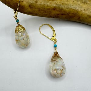 May include: A pair of gold-coloured drop earrings with teardrop-shaped pendants. The pendants are filled with iridescent, white, and gold-coloured fragments. Each earring has a small turquoise bead and a decorative gold cap.
