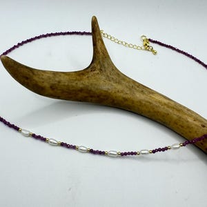 May include: A delicate necklace featuring small, faceted deep purple beads interspersed with white, oval-shaped pearls and tiny gold-toned metallic spheres. The necklace includes a gold-toned chain extender with a lobster claw clasp closure.