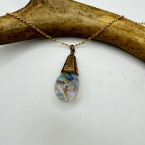 May include: A gold-colored necklace with a teardrop-shaped pendant. The pendant has a gold-colored cap and contains iridescent, pearlescent material. The necklace is displayed on a light brown antler against a white background.