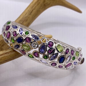 May include: A silver bangle bracelet with a mosaic of colorful gemstones. The bracelet features a variety of shapes and colors, including purple, blue, green, and yellow stones. The bracelet is set against a light background.