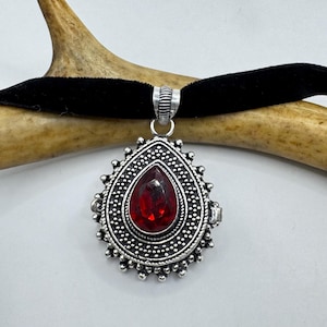 May include: A silver pendant necklace with a red teardrop-shaped stone set in a detailed silver frame. The necklace is on a black velvet choker.