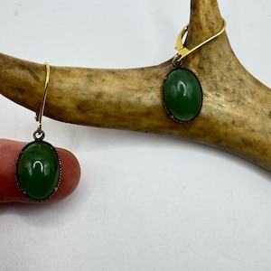 Op de afbeelding: Een paar ovale oorbellen met groene stenen en goudkleurige haken. De oorbellen zijn gezet in een donker metalen frame en worden op een lichte achtergrond getoond. Een klassiek sieraad.