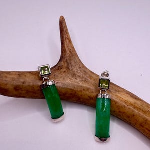 Op de afbeelding: Een paar oorhangers met een heldergroene, cilindrische jadesteen met een vierkante, lichtgroene edelsteen aan de bovenkant. De oorbellen hebben zilverkleurige metalen accenten en worden gepresenteerd op een lichtbruin gewei.