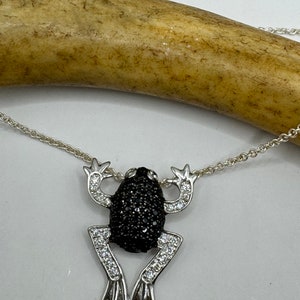 May include: A silver frog pendant necklace. The frog's body is black, and its legs and arms are adorned with clear crystals. The necklace chain is also silver. The frog is posed as if it is jumping.