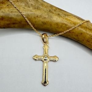 Può includere: Una collana con pendente a forma di croce color oro. La croce presenta una piccola gemma trasparente al centro e dettagli ornati. Anche la catena è color oro. La collana è esposta su uno sfondo neutro.