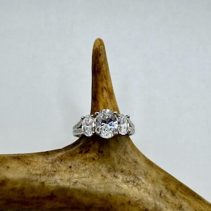 May include: A silver ring featuring three oval-shaped clear stones. The ring is displayed on a light brown antler.