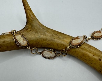 May include: A gold chain bracelet with four oval-shaped cameo pendants. Each cameo features a white cameo profile with a brown background.