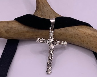May include: A silver crucifix pendant with a thorny design and a figure of Jesus Christ. The pendant is hanging from a black velvet choker.
