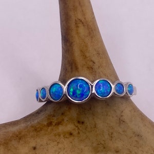 Può includere: Un anello d'argento con una serie di pietre di opale blu rotonde. Gli opali hanno una qualità vibrante e iridescente, con lampi di verde e blu. L'anello è posto su uno sfondo neutro.