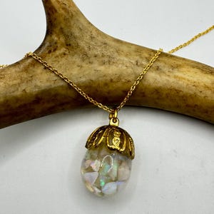 May include: A gold-colored necklace with a pendant featuring a gold cap and a clear, iridescent stone. The necklace is displayed on a light brown antler against a white background. The pendant is approximately 1 inch in length.
