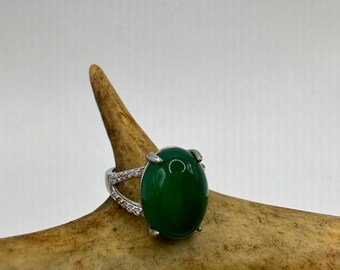 May include: A silver ring featuring a large, oval, deep green gemstone. The band splits on either side of the stone, with one side adorned with small, clear accent stones. The ring is displayed on a light brown antler against a white background.