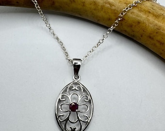 May include: A silver necklace with an oval pendant featuring a floral filigree design. The pendant has a small, round, red gemstone at its center. The necklace chain is delicate and silver, and the background is a light color.