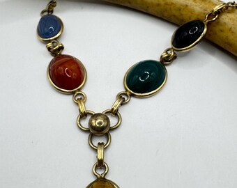 Puede incluir: Collar vintage dorado con una serie de piedras ovaladas y redondas de colores. El collar tiene un colgante central con una piedra ovalada marrón. Las piedras son en tonos azul, rojo, verde y negro.