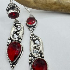 May include: A pair of silver-toned drop earrings with large, faceted, red gemstones. The earrings feature an ornate, swirling design and are suspended from a simple hook. The gemstones are teardrop-shaped and oval-shaped.