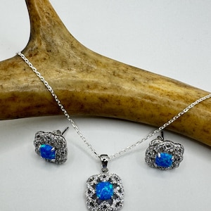 May include: A silver necklace and earring set featuring square, blue opal stones. The pendant and earrings are surrounded by a halo of small, clear stones. The jewelry is displayed on a white surface with a brown antler in the background.