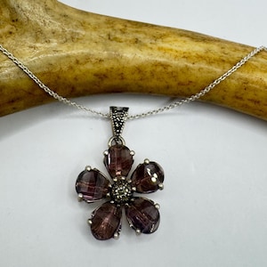 May include: A silver necklace featuring a flower pendant with five dark purple, teardrop-shaped stones. The center of the flower has a cluster of smaller, silver-toned stones. The chain is delicate and silver.