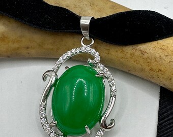 May include: A green oval gemstone pendant necklace with a silver-coloured frame, accented with small clear stones. The pendant hangs from a black velvet choker.
