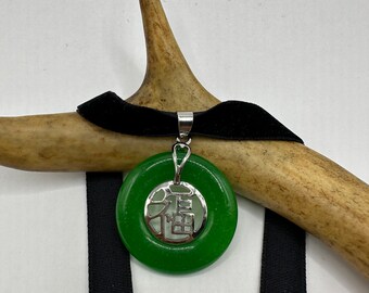May include: A green jade pendant with a silver charm featuring the Chinese character for "good fortune" hanging from a black velvet choker.
