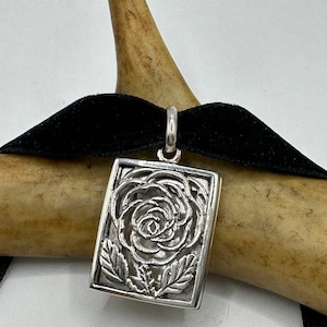 May include: A silver pendant featuring a detailed cutout design of a rose with leaves. The pendant is rectangular in shape and hangs from a black velvet ribbon.