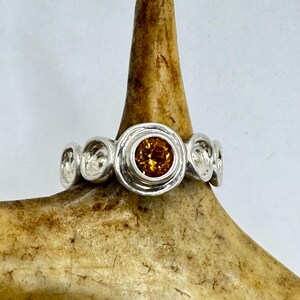 May include: A silver ring with a round, orange gemstone set in a textured band. The ring is displayed on a brown antler.