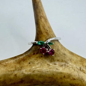 May include: A silver ring featuring a cherry design. The ring has two red gemstone cherries with green leaves. The ring is displayed on a light brown antler against a white background.
