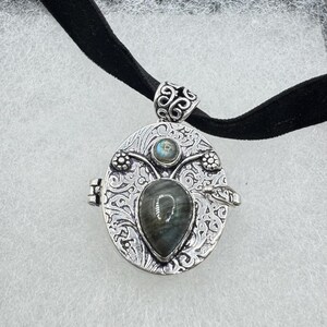 Puede incluir: Un collar de plata con un dije de medall&oacute;n adornado con una piedra de labradorita en forma de coraz&oacute;n y una piedra de labradorita redonda m&aacute;s peque&ntilde;a. El medall&oacute;n tiene un dise&ntilde;o floral intrincado y una abertura con bisagra.