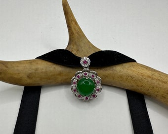 May include: A green jade pendant with a silver setting and pink gemstones. The pendant is hanging from a black velvet ribbon.