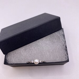 May include: A silver ring with a single pearl and small clear stones set in a delicate design. The ring is displayed in a black velvet box with white padding.