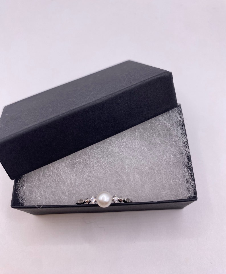 May include: A silver ring with a single pearl and small clear stones set in a delicate design. The ring is displayed in a black velvet box with white padding.