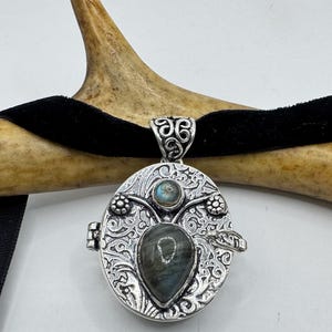 Puede incluir: Un colgante de plata en forma de medall&oacute;n con una abertura con bisagra, adornado con un dise&ntilde;o floral decorativo y dos piedras de labradorita iridiscentes.