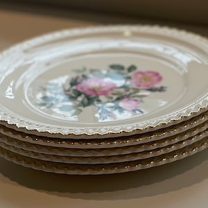 Pode incluir: Uma pilha de cinco pratos de cerâmica branca com uma borda dourada e um design floral. Os pratos apresentam uma rosa rosa com folhas verdes.
