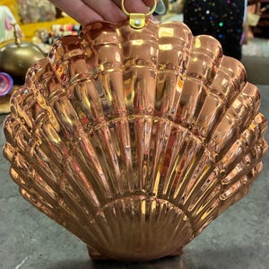 May include: A shiny copper-colored metal mold shaped like a large scallop shell. The mold features a series of vertical ridges radiating from the base to the scalloped edge. A small circular metal ring is attached to the top for hanging purposes.
