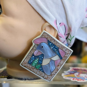 May include: A square earring with a mosaic design featuring a blue bird with pink and white accents. The earring is set in a speckled frame. The earring is attached to a mannequin head wearing a floral patterned headscarf.
