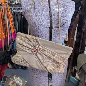 Puede incluir: Un bolso de mano vintage beige con una correa larga, exhibido en un maniqu&iacute;. El bolso presenta una solapa texturizada con un cierre decorativo. El fondo incluye percheros y otros accesorios.