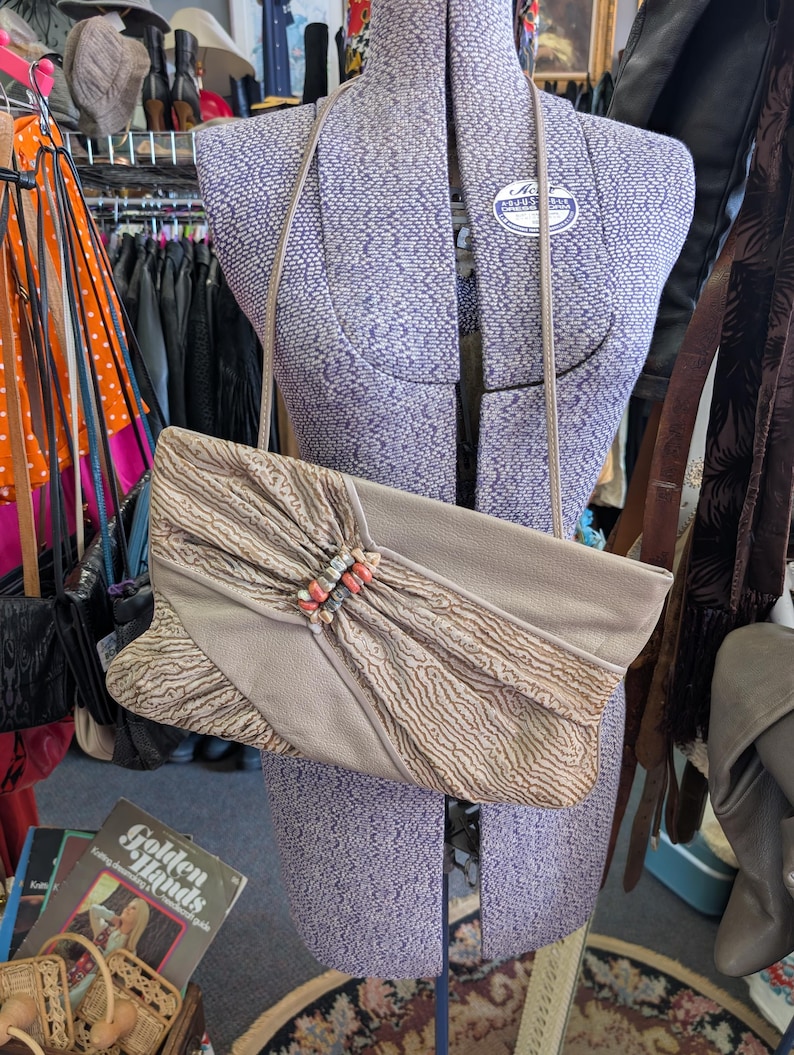 Puede incluir: Un bolso de mano vintage beige con una correa larga, exhibido en un maniqu&iacute;. El bolso presenta una solapa texturizada con un cierre decorativo. El fondo incluye percheros y otros accesorios.