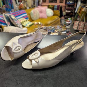 May include: A pair of off-white leather slingback sandals with a peep-toe design. Each sandal features a decorative oval buckle and a low heel. The shoes are displayed on a dark surface, showcasing a vintage style.