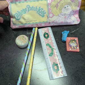May include: A pink vinyl pencil case with a Cabbage Patch Kids logo and a picture of a Cabbage Patch Kid doll. The case is open and contains a ruler, two pencils, an eraser, and a sticker.