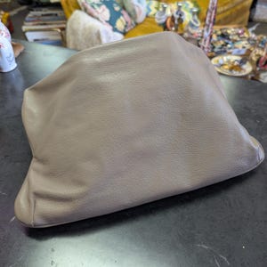 Puede incluir: Un bolso de mano de color topo con un aspecto suave y texturizado. El bolso est&aacute; hecho de un material flexible y tiene un dise&ntilde;o fruncido. El bolso de mano est&aacute; sobre una superficie oscura y reflectante.