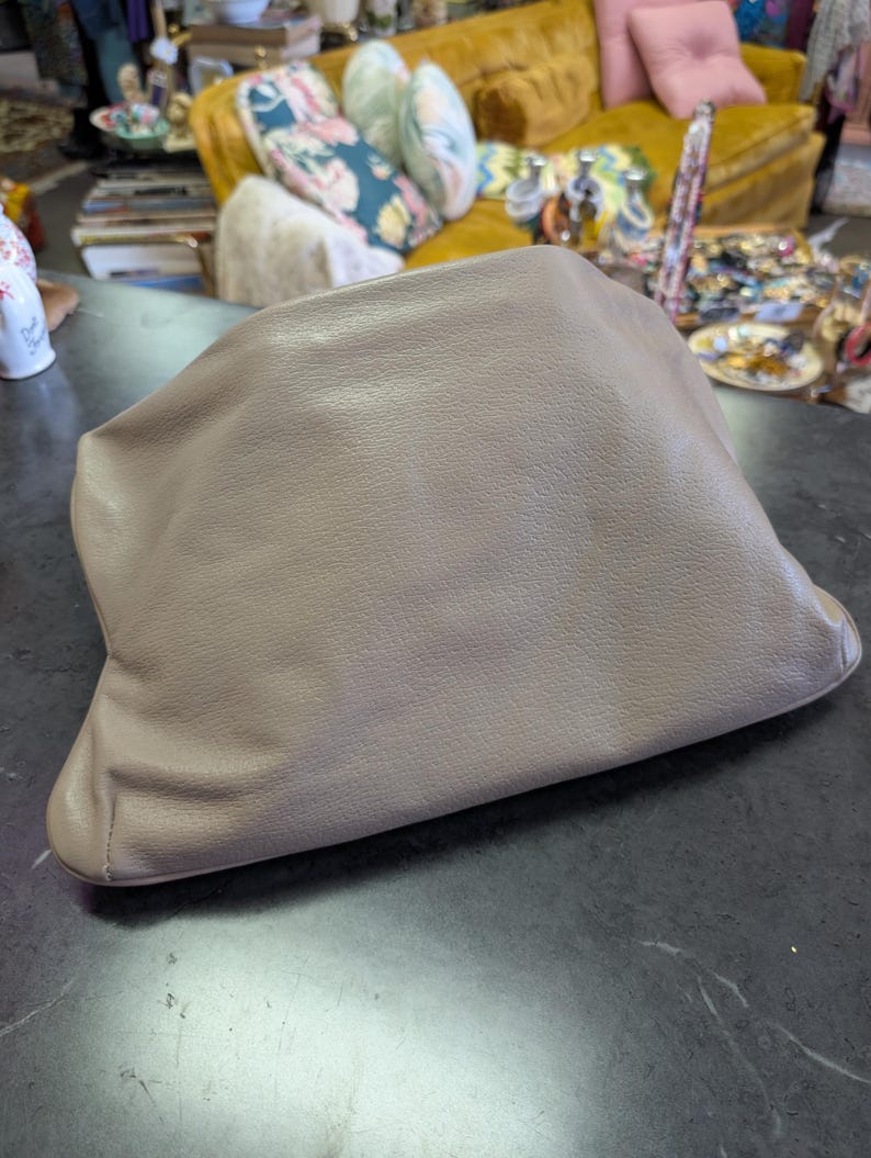 Puede incluir: Un bolso de mano de color topo con un aspecto suave y texturizado. El bolso est&aacute; hecho de un material flexible y tiene un dise&ntilde;o fruncido. El bolso de mano est&aacute; sobre una superficie oscura y reflectante.